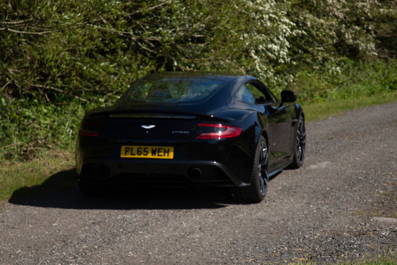 The rear of a black Aston Martin Vanquish on a country lane, yellow number plate visible
