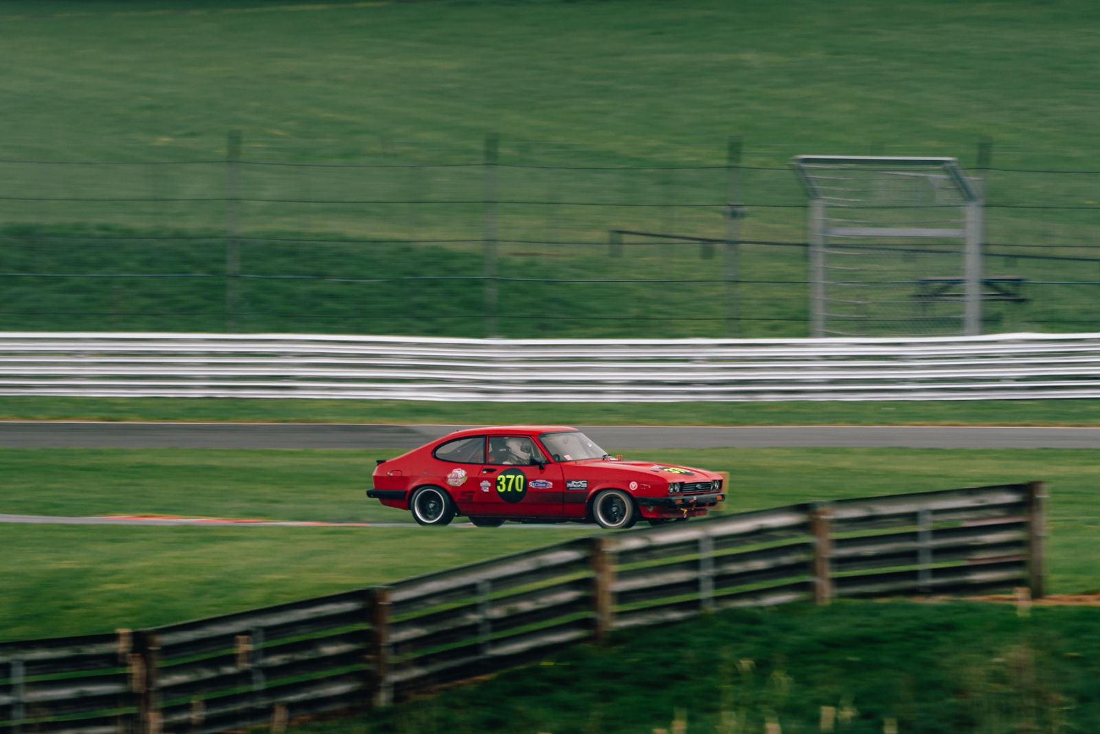 A red Ford Capri race car panned against green grass at Oulton Park